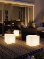 Three lit white Clérachic cube lamps in different sizes on a timber deck at dusk, in front of a glazed living room.