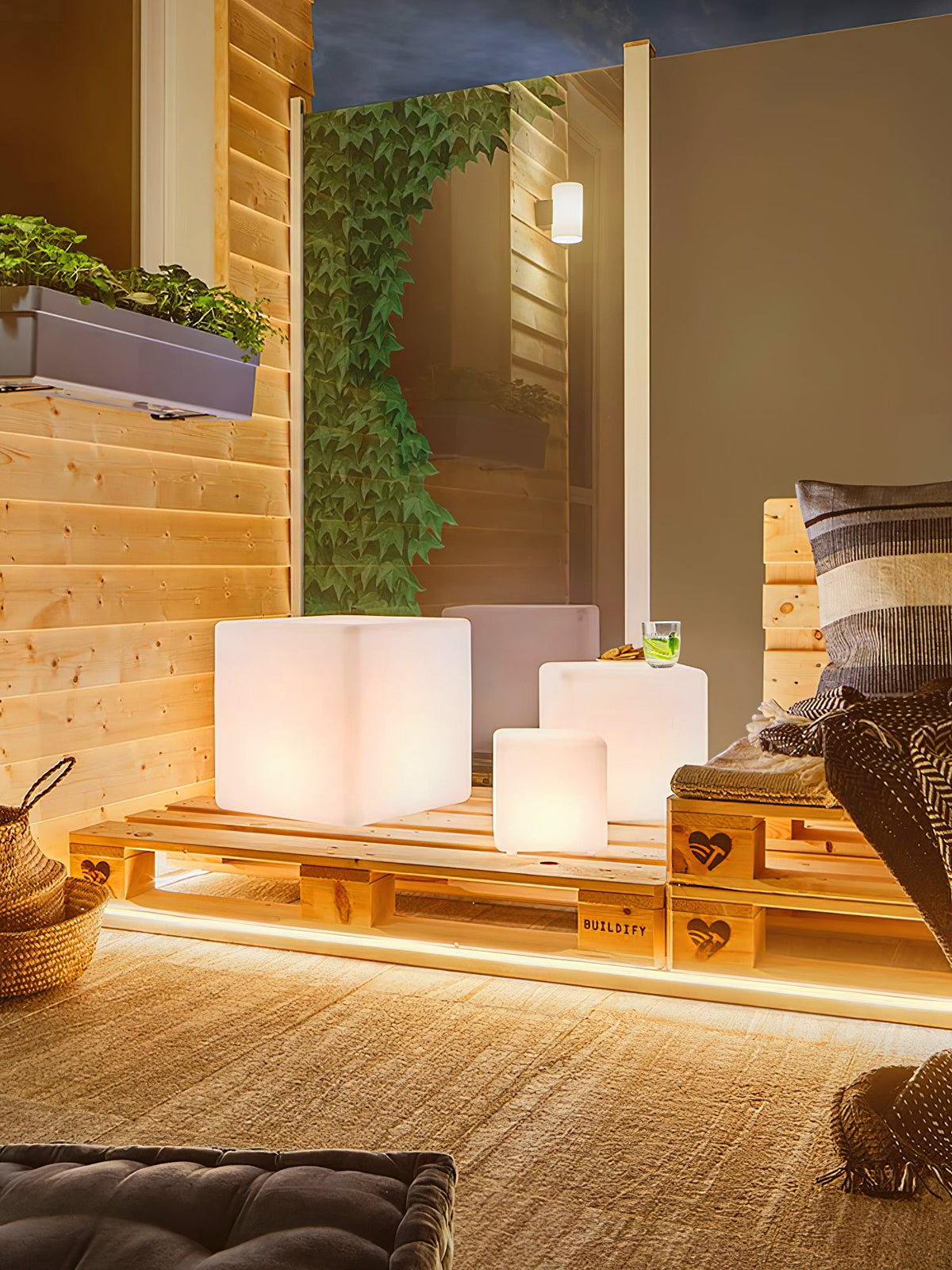 A lit white Clérachic cube lamp on a timber patio, beside a pallet seat, a climbing plant and two smaller cubes.