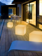 Three lit white Clérachic cube lamps of different sizes on a timber deck beside a lounger and a glass-fronted house.