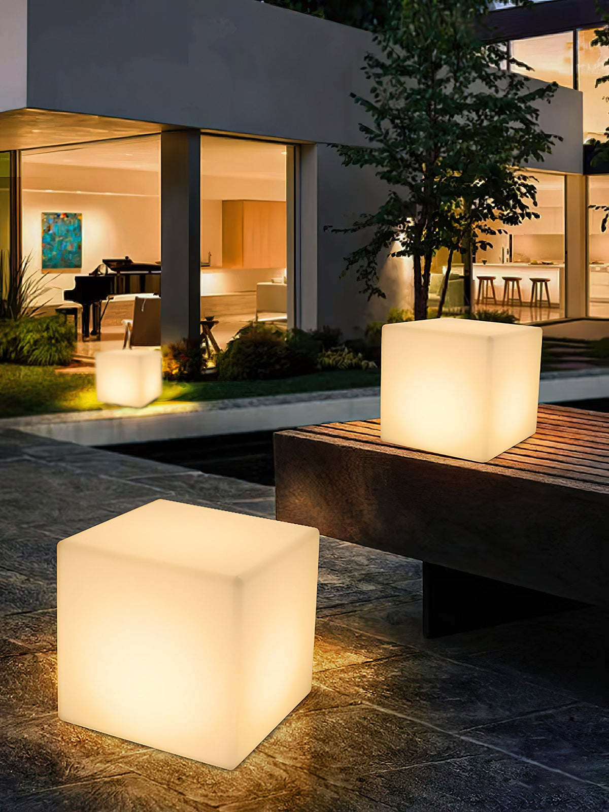 Two lit white Clérachic cube lamps on a paved terrace at dusk, one on a stone bench, in front of a lit house.