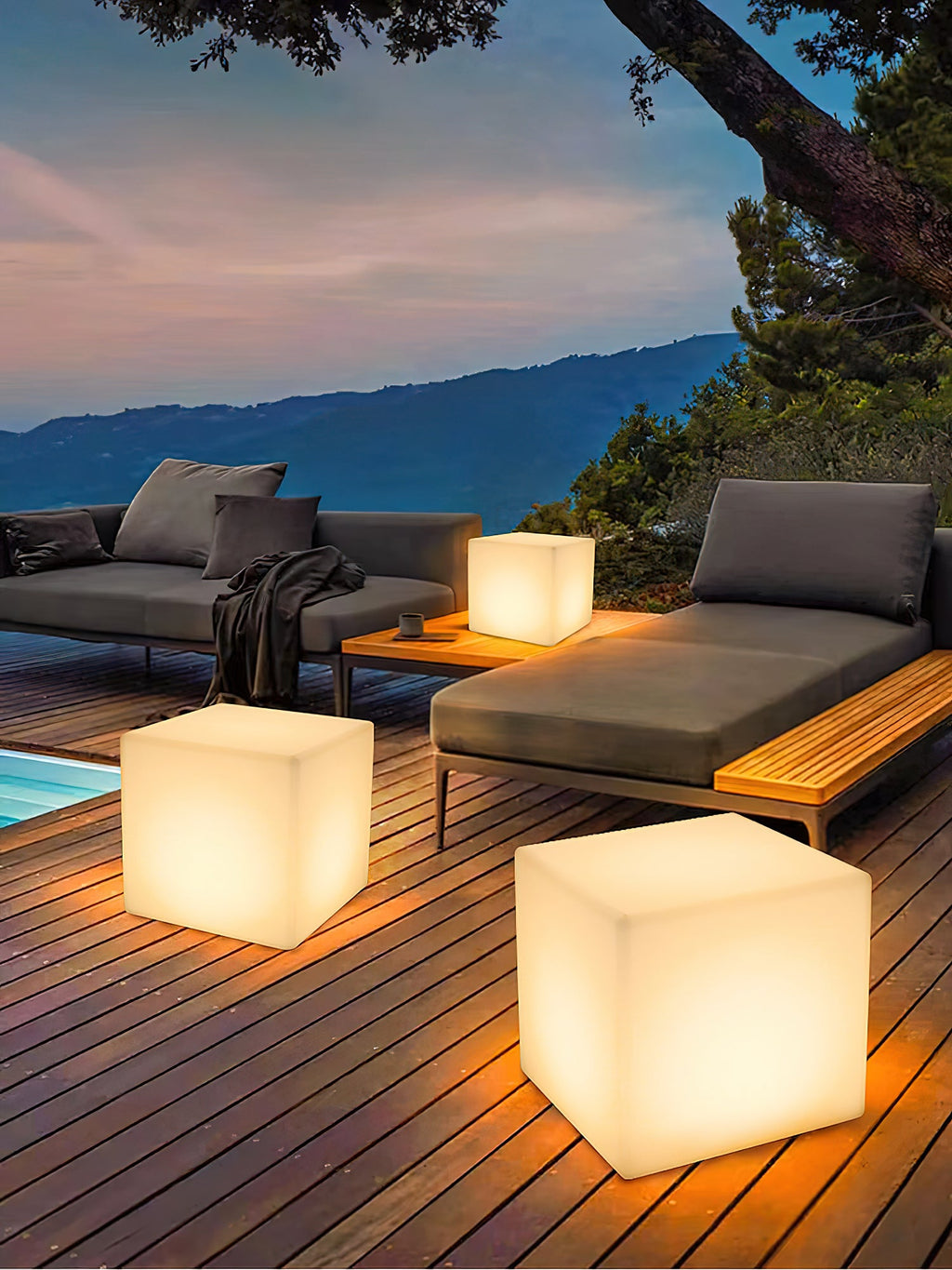 Three lit white Clérachic cube lamps on a timber deck beside a grey outdoor sofa, with a mountain view at dusk.