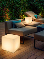 On a timber deck behind a grey outdoor sofa, one white Clérachic cube lamp glows, with another cube on a side table.