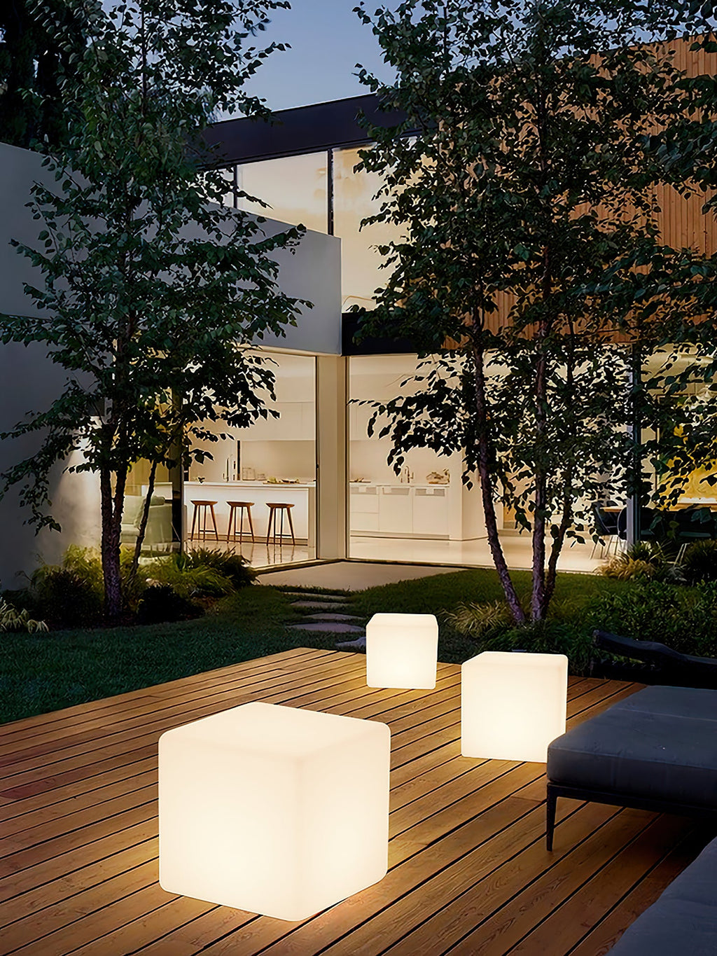 Three lit white Clérachic cube lamps of different sizes on a garden deck at dusk, with trees and a lit kitchen behind.
