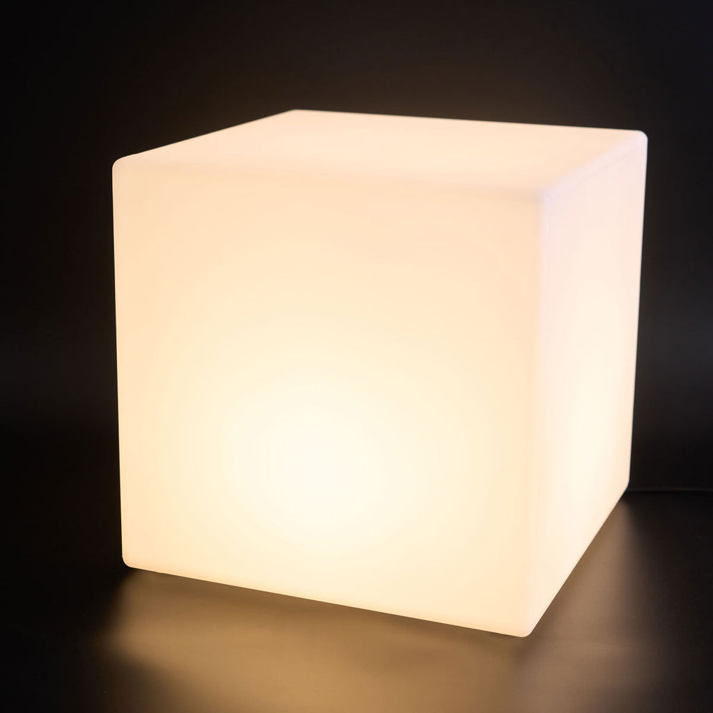 A large lit white Clérachic cube lamp with a warm glow and rounded corners, on a black backdrop.
