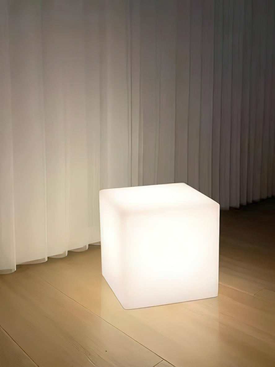 A white Clérachic cube lamp lit cool white on a wooden floor, in front of floor-length sheer curtains.