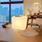 Beside a wooden side table with a floor lamp and reed diffuser, this white Clérachic cube lamp glows, with a cat on a rug.