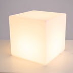 Front view of a lit white Clérachic cube lamp with a warm glow, on a pale surface against a grey wall.