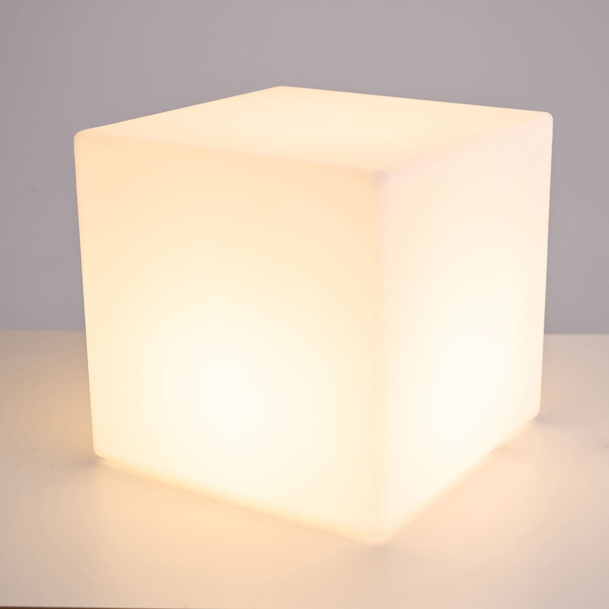 Front view of a lit white Clérachic cube lamp with a warm glow, on a pale surface against a grey wall.