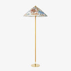 This Clérachic floor lamp on a plain white backdrop, with a wide conical shade printed with a colourful floral pattern on a slim brass stem.