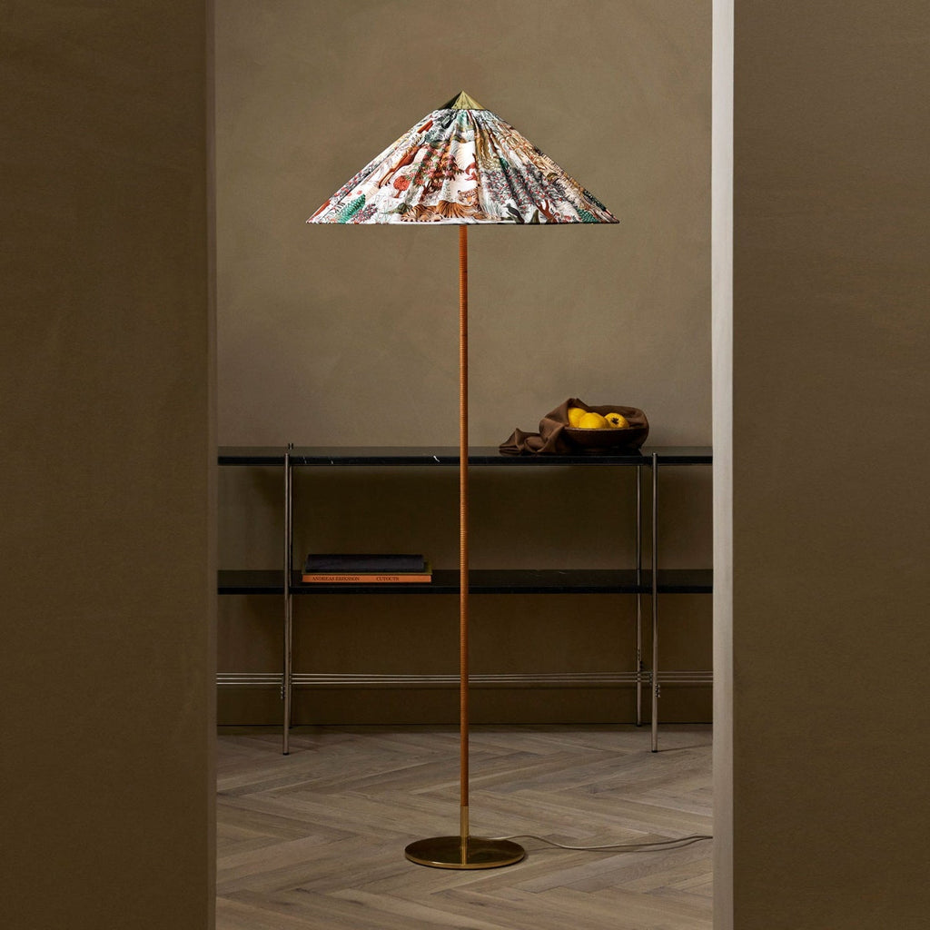 A Clérachic floor lamp with a floral printed conical shade stands in a doorway, beside a black metal shelf, on a herringbone wood floor.