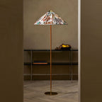 A Clérachic floor lamp with a floral printed conical shade stands in a doorway, beside a black metal shelf, on a herringbone wood floor.