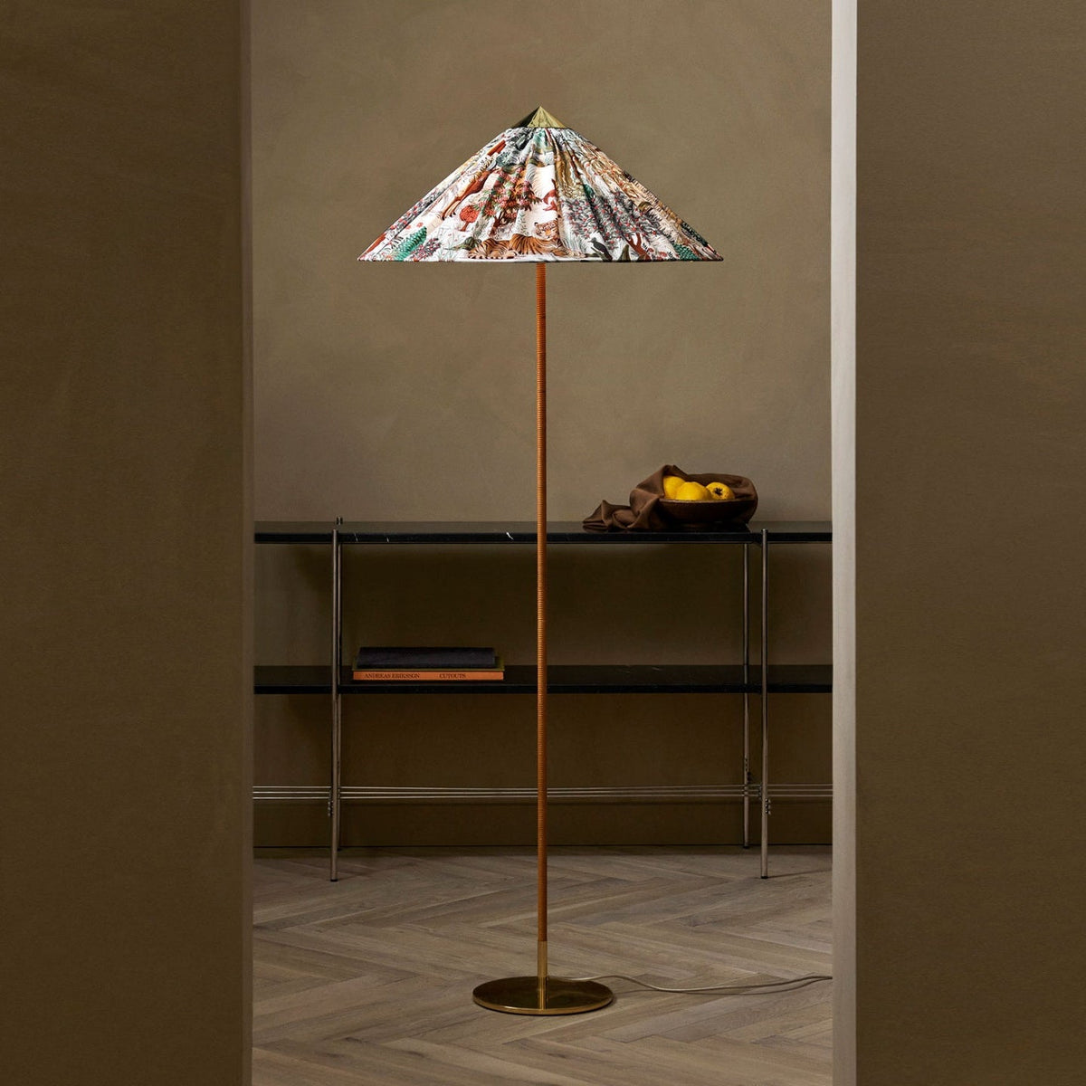 A Clérachic floor lamp with a floral printed conical shade stands in a doorway, beside a black metal shelf, on a herringbone wood floor.