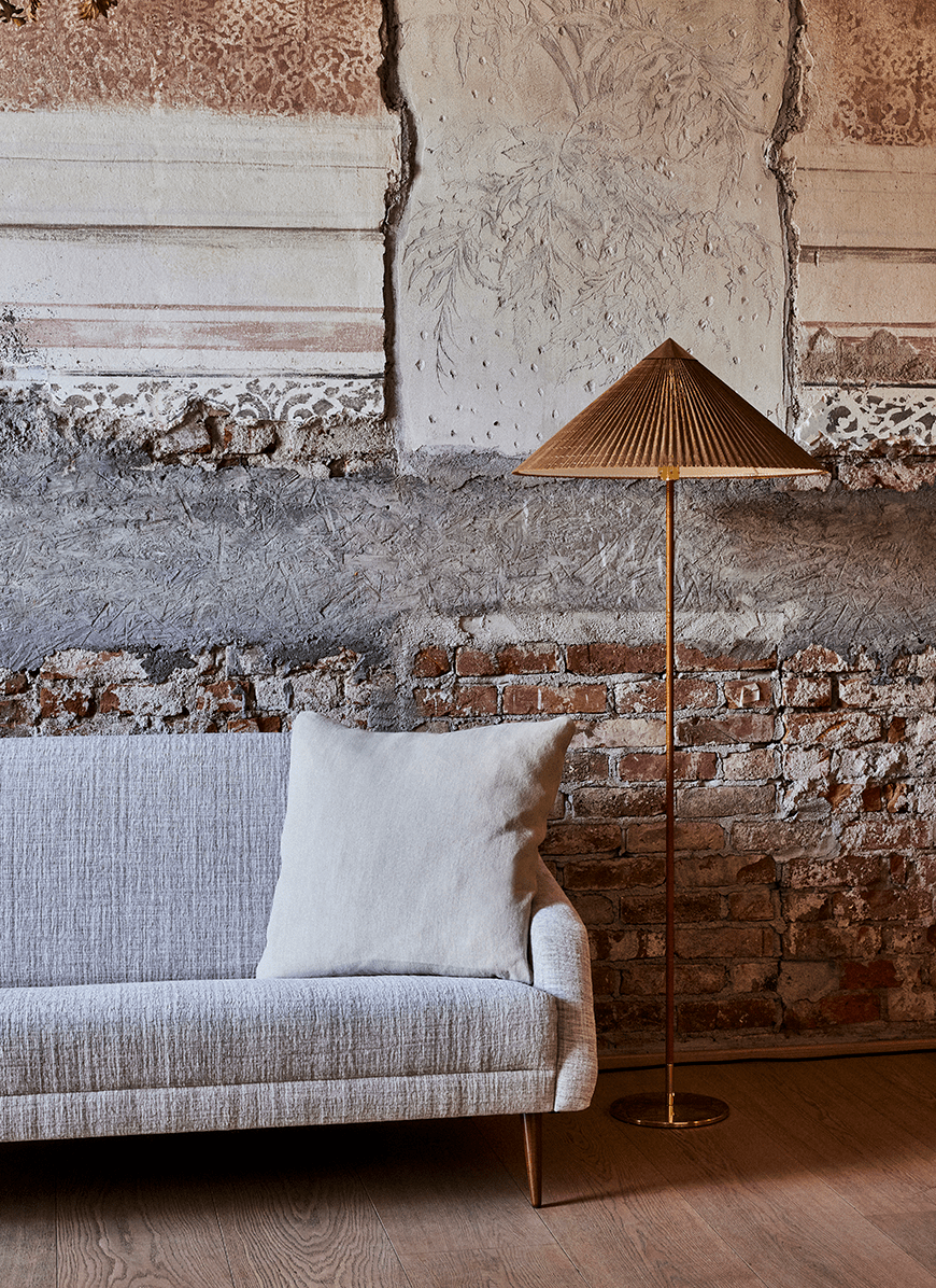 This Clérachic floor lamp with a woven natural shade stands against a peeling plaster and brick wall, beside a grey sofa with a cushion.