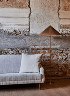 This Clérachic floor lamp with a woven natural shade stands against a peeling plaster and brick wall, beside a grey sofa with a cushion.