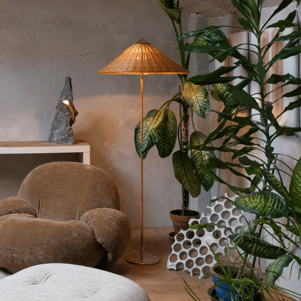 This Clérachic floor lamp with a woven rattan shade stands among large leafy plants and a velvet armchair, in front of a plaster wall.