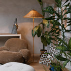 This Clérachic floor lamp with a woven rattan shade stands among large leafy plants and a velvet armchair, in front of a plaster wall.