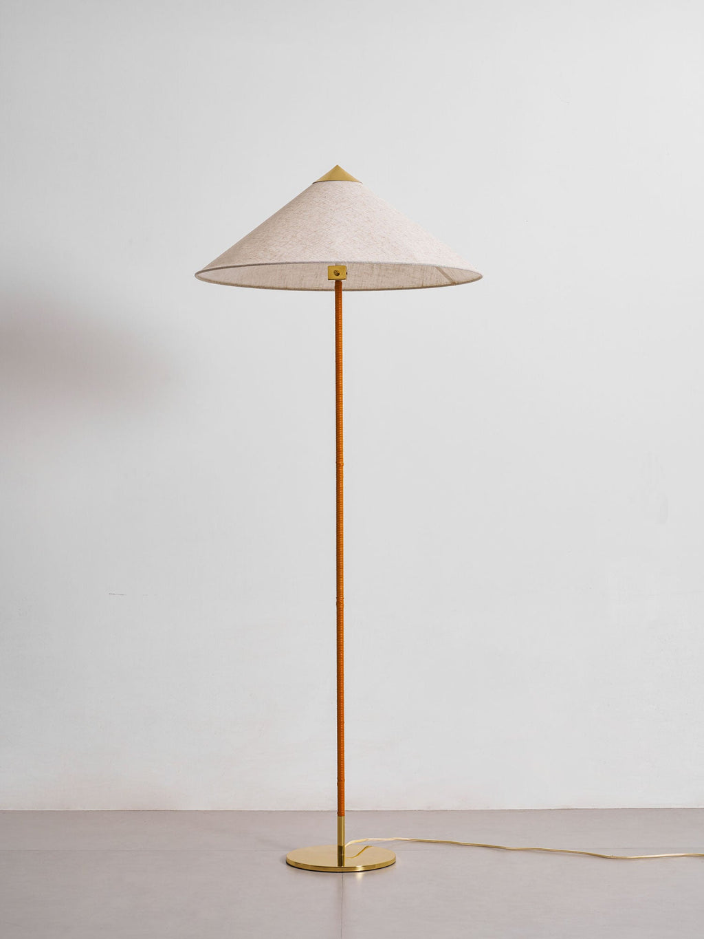 In this photo, a Clérachic floor lamp with a natural fabric shade and a wrapped stem on a round brass base, seen at a lower angle against a grey wall.