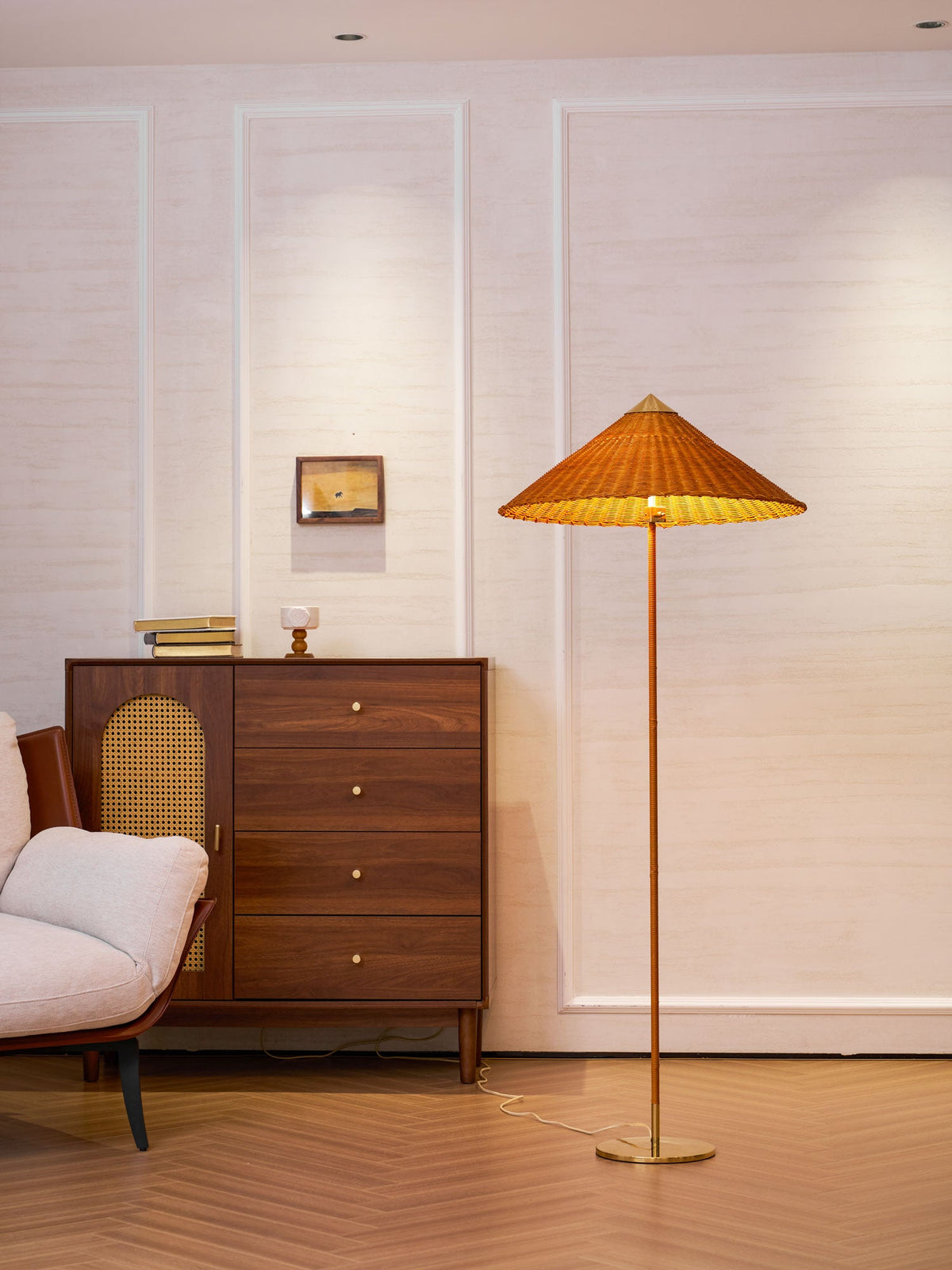Lit Clérachic floor lamp with a woven rattan shade, beside a walnut chest of drawers with a cane door, on a herringbone floor.