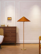 A Clérachic floor lamp with a glowing rattan shade stands next to a dark wood chest of drawers, against a white panelled wall.