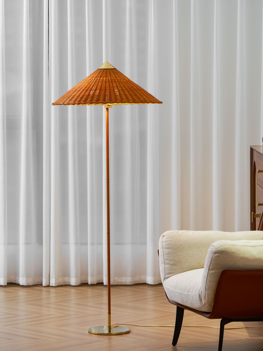 This Clérachic floor lamp with a rattan shade and a brass base stands against sheer curtains, with a cream armchair at the edge of the frame.