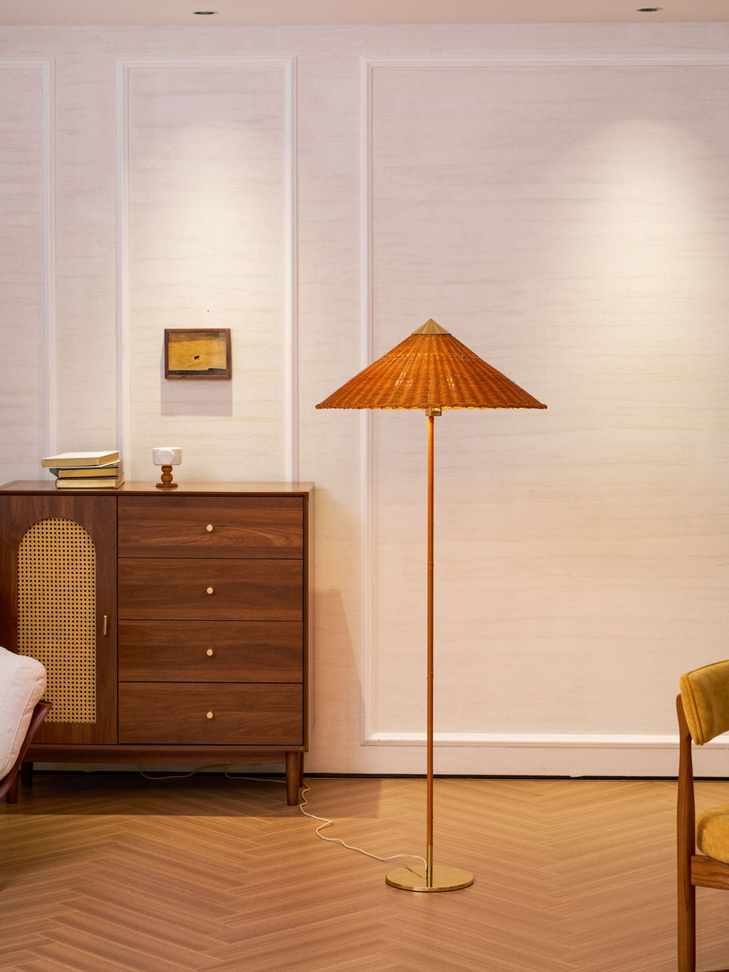 Clérachic floor lamp with a warm rattan shade stands by a cane-fronted walnut chest, with a mustard chair at the edge of the frame.