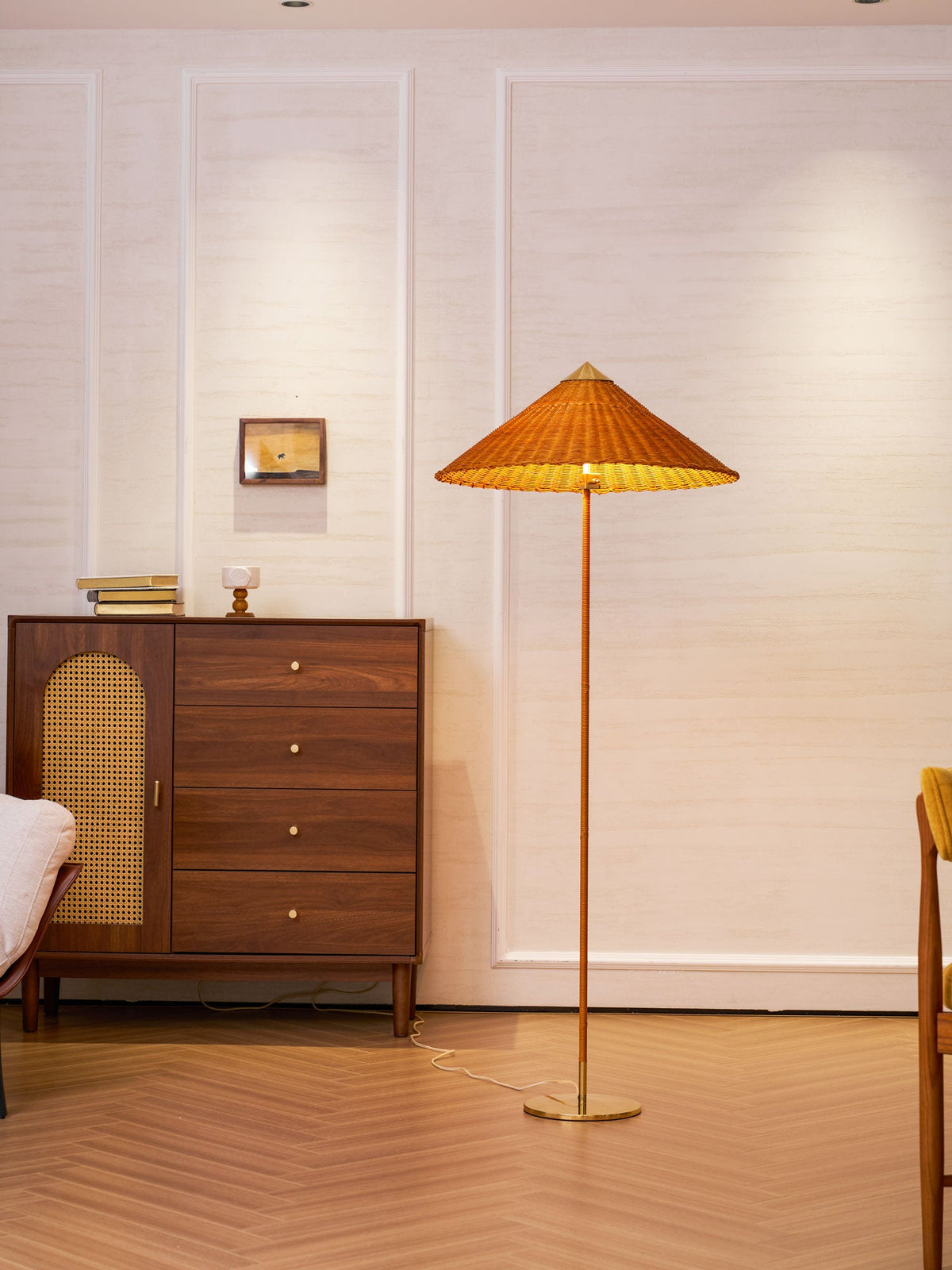Shown here, a Clérachic floor lamp with a rattan shade casts a warm glow on a panelled wall, next to a walnut chest and a chair upholstered in mustard.