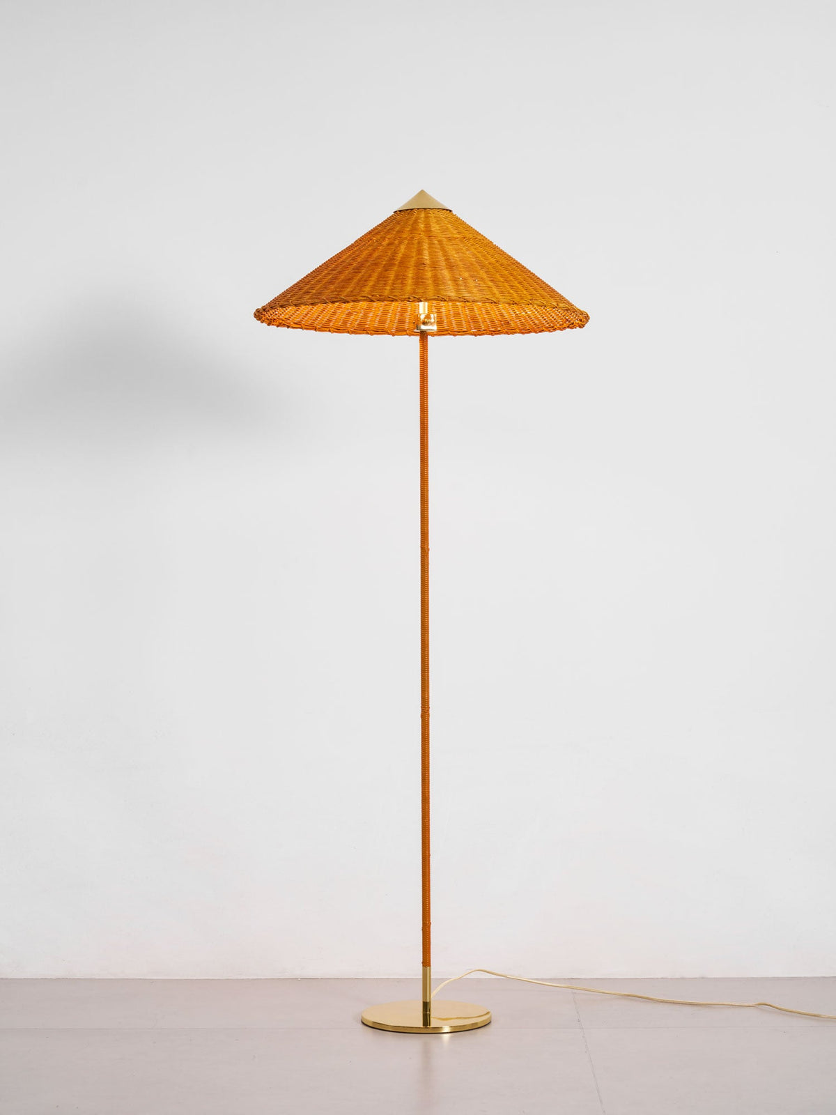 In this photo, a Clérachic floor lamp with a woven rattan shade and a brass base on a pale floor, against a plain white wall.