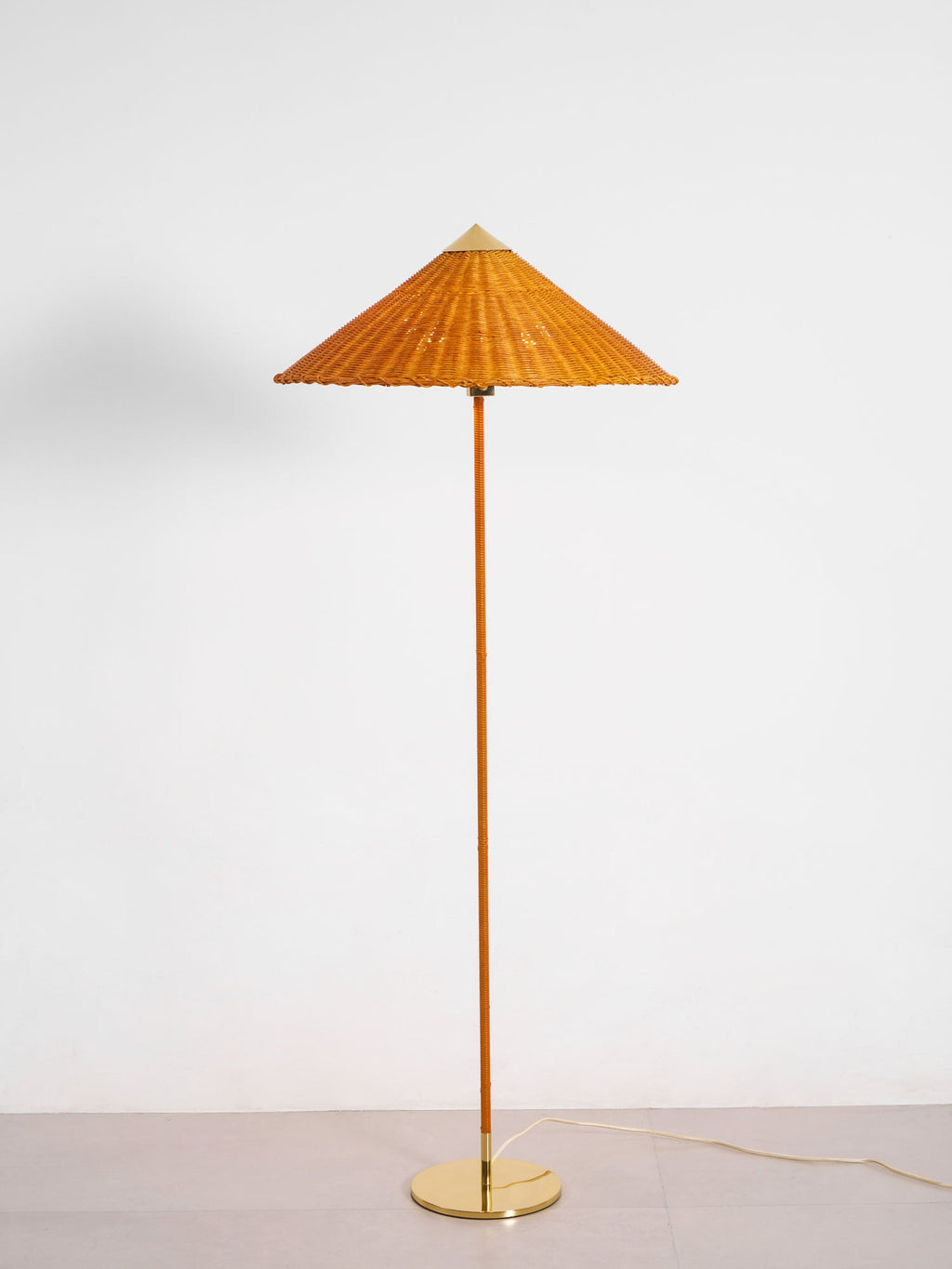 This Clérachic floor lamp with a rattan shade and a cane-wrapped stem stands on a brass base, shown against a light wall and floor.