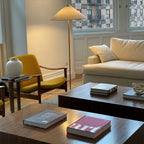 This Clérachic floor lamp with a pale fabric shade stands in a living room with a mustard armchair, a cream sofa and a low wood table.