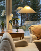 Pictured here, a Clérachic floor lamp with a beige fabric shade beside a large window, over an armchair with a tan blanket and an orchid on a table.