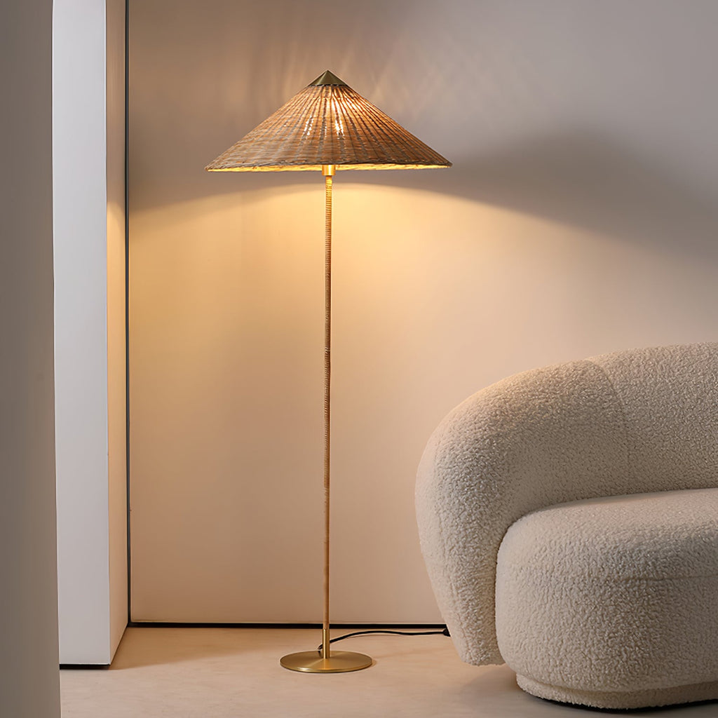 This Clérachic floor lamp with a natural rattan shade stands lit against a cream wall, beside a boucle armchair, its brass base on the floor.