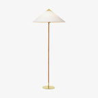 Front view of this Clérachic floor lamp with a white conical shade, a rattan-wrapped stem and a round brass base on a plain backdrop.