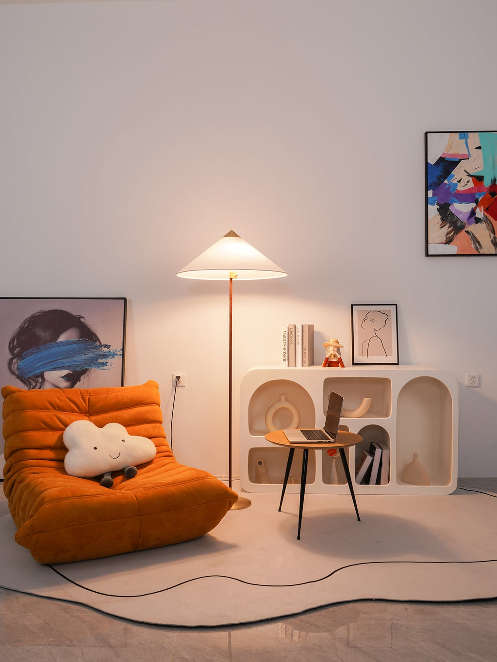 A Clérachic floor lamp with a white shade lights an orange lounge chair, a low white shelf and a small desk, in a room with a rounded rug.