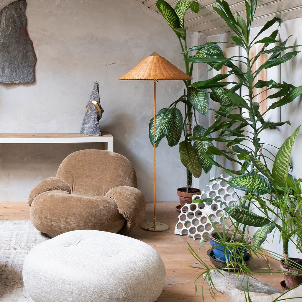 Clérachic floor lamp with a rattan shade among tall green plants, beside a velvet armchair and a round white pouffe.