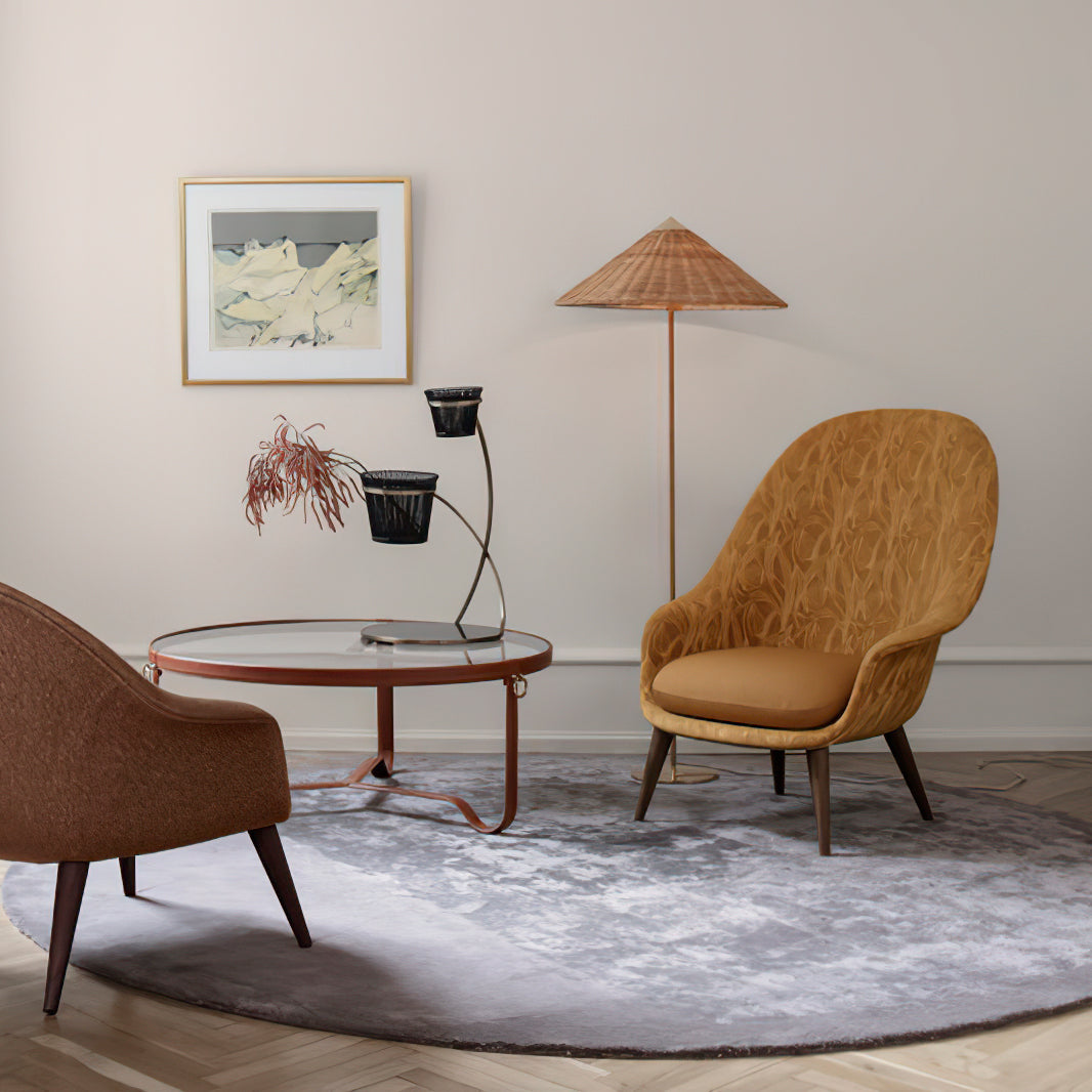 Pictured here, a Clérachic floor lamp with a woven shade stands behind a round glass coffee table, between an ochre chair and a rust-brown one.