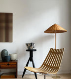 This Clérachic floor lamp with a woven rattan shade stands next to a slatted wooden chair and a small black stool, by a white wall.