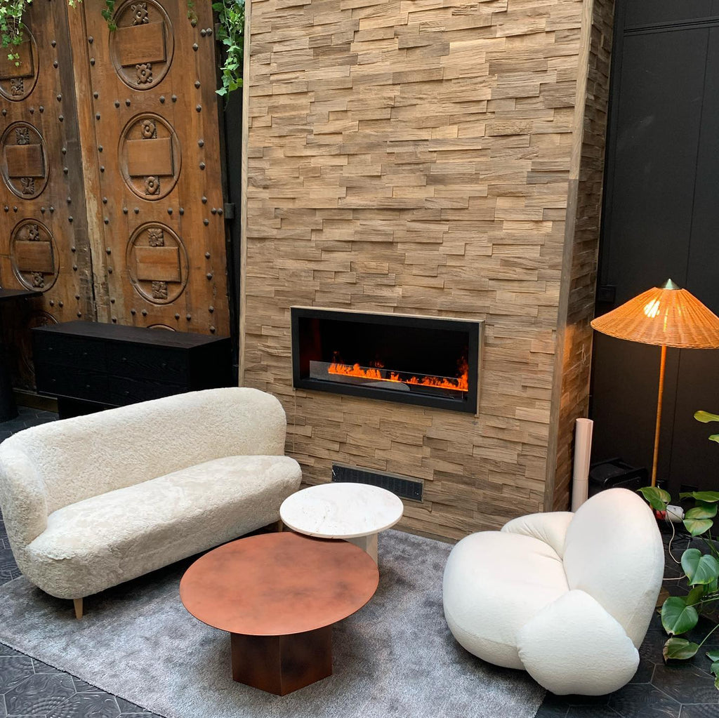 Shown here, a Clérachic floor lamp with an orange-toned shade in a corner by a stone-clad fireplace, with a white sofa and two low round tables.