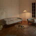 This Clérachic floor lamp with a white shade stands behind a pale curved sofa, near a glass coffee table, on a herringbone floor.