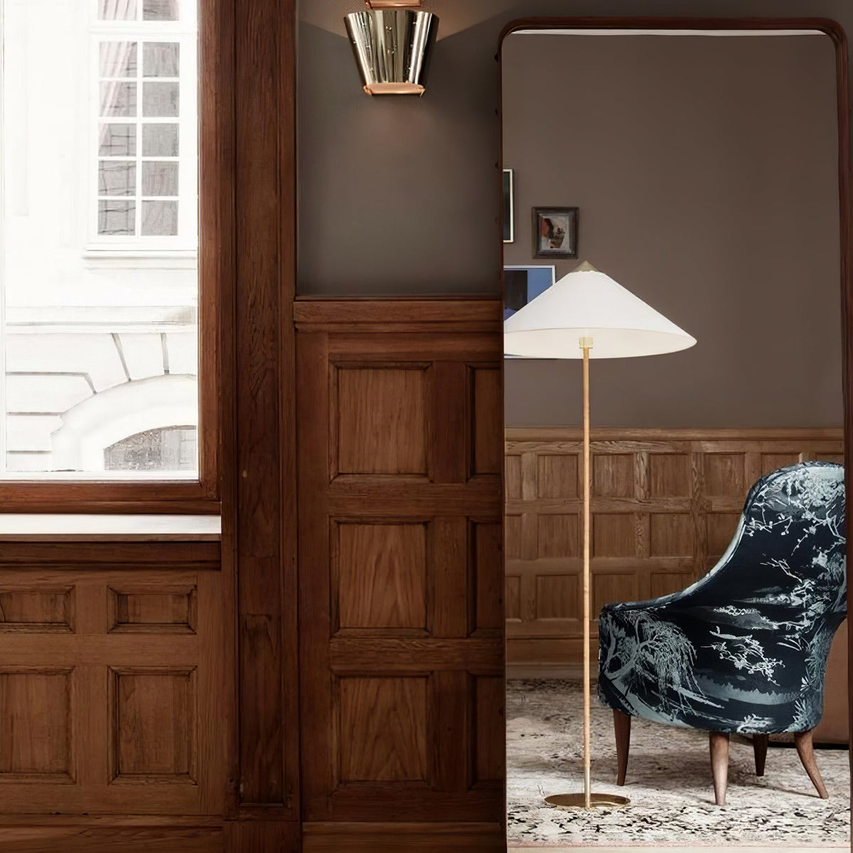 In this photo, a Clérachic floor lamp with a white conical shade stands next to a dark wood wall, with an armchair upholstered in blue floral fabric.