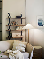 In this photo, a Clérachic floor lamp with a white conical shade stands behind a cream sofa, beside wall shelves and a framed blue wave print.
