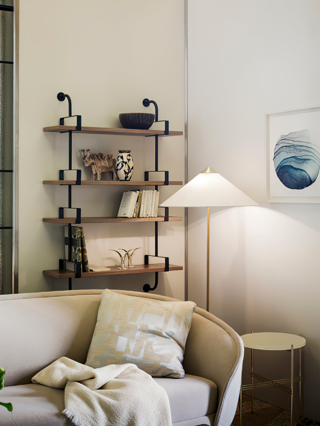 In this photo, a Clérachic floor lamp with a white conical shade stands behind a cream sofa, beside wall shelves and a framed blue wave print.