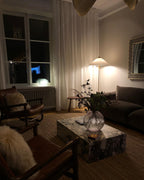 Clérachic floor lamp with a small pale shade glowing in a dark living room, with a marble coffee table, leather chairs and a glass vase.