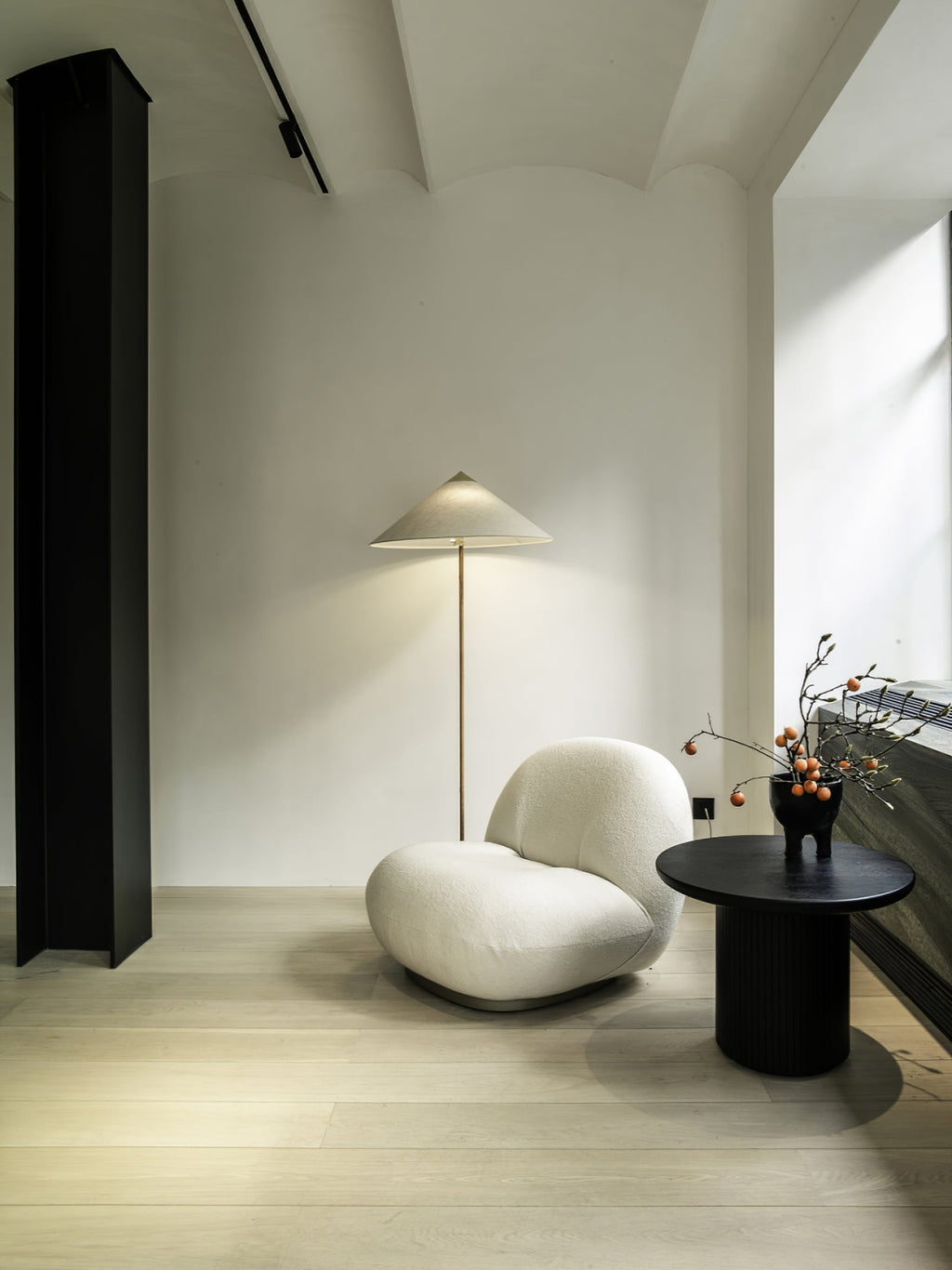 This Clérachic floor lamp with a pale conical shade stands in a bright, minimal room, beside a white rounded armchair and a black side table.