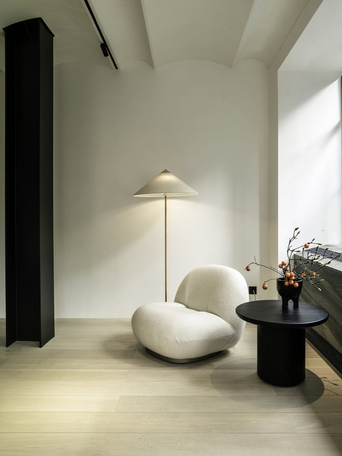This Clérachic floor lamp with a pale conical shade stands in a bright, minimal room, beside a white rounded armchair and a black side table.