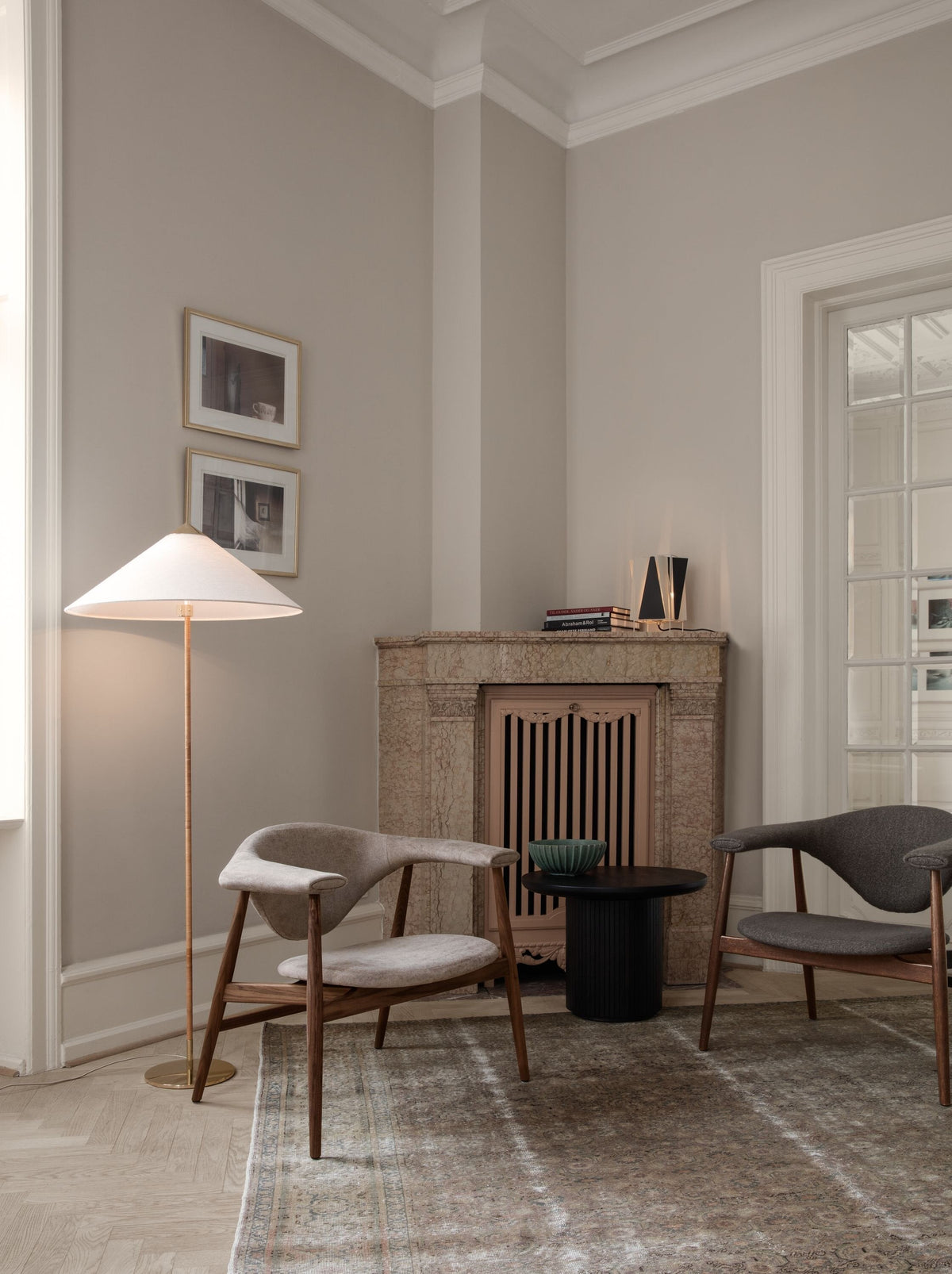Pictured here, a Clérachic floor lamp with a white shade stands in a corner with a stone fireplace, near two upholstered armchairs and a worn rug.