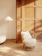 A Clérachic floor lamp with a white shade stands beside a rattan armchair with a white cushion, in front of oak panelling and a herringbone floor.