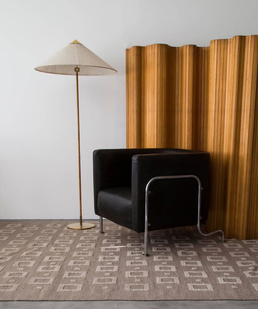This Clérachic floor lamp with a pale woven shade stands on a patterned rug, beside a black leather armchair and a wooden folding screen.