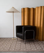 This Clérachic floor lamp with a pale woven shade stands on a patterned rug, beside a black leather armchair and a wooden folding screen.