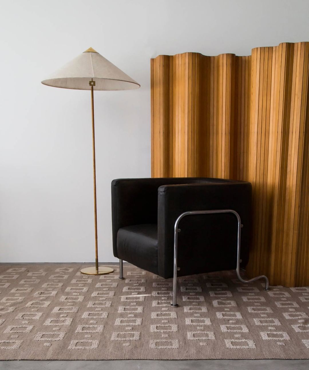 This Clérachic floor lamp with a pale woven shade stands on a patterned rug, beside a black leather armchair and a wooden folding screen.