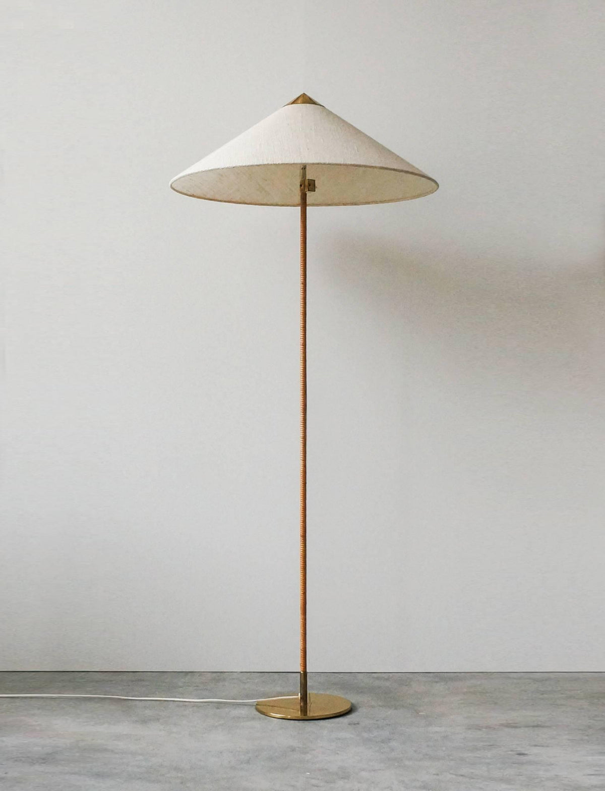 This Clérachic floor lamp with a pale textured shade and a rattan-wrapped stem stands against a grey wall on a concrete floor.